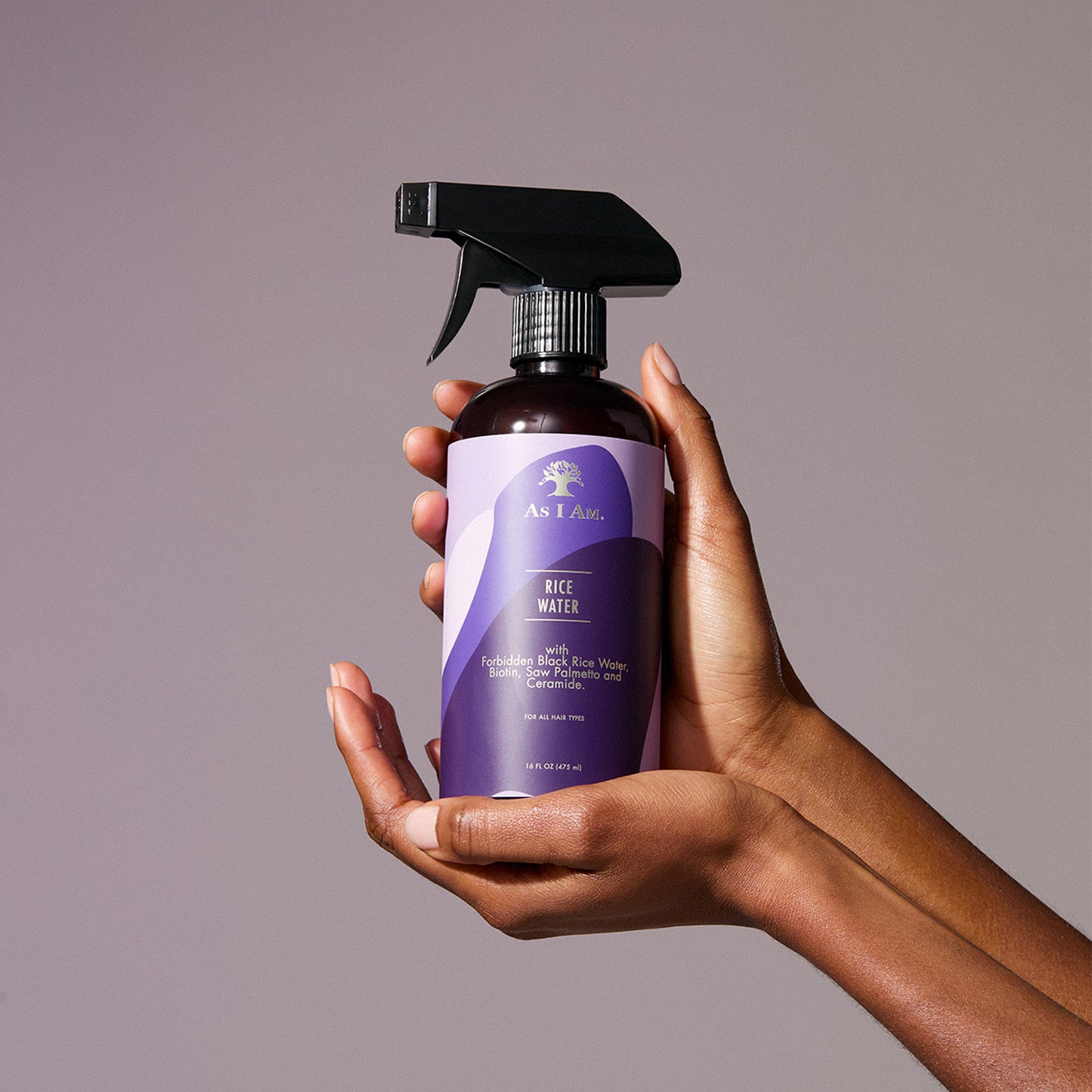 Una mano sosteniendo un frasco con atomizador del spray para el crecimiento del cabello «As I Am Rice Water» con una etiqueta morada.