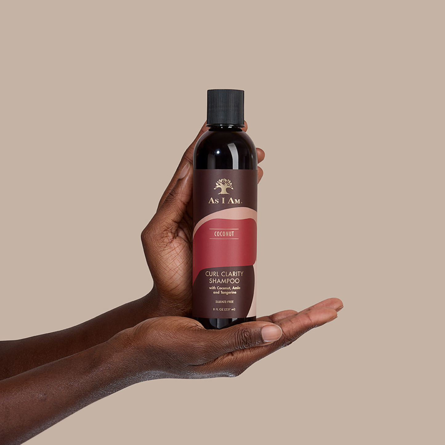 Mano sosteniendo una botella de champú As I Am Coconut Curl Clarity sobre un fondo beige.