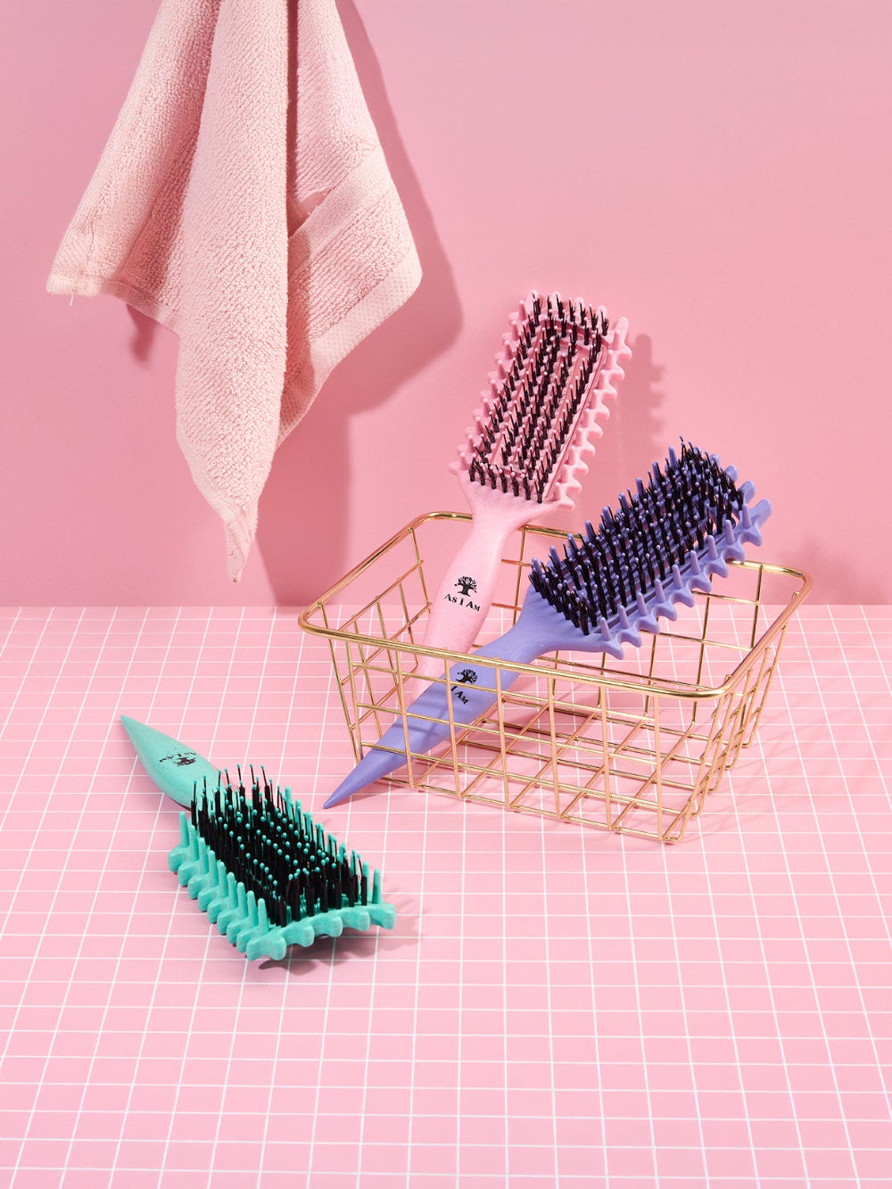 Tres cepillos para el pelo de distintos colores sobre un fondo cuadriculado rosa con una cesta dorada.