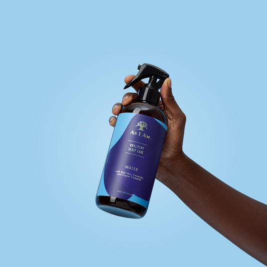 Mano sosteniendo un frasco de desodorante en spray AS I AM sobre un fondo azul claro.